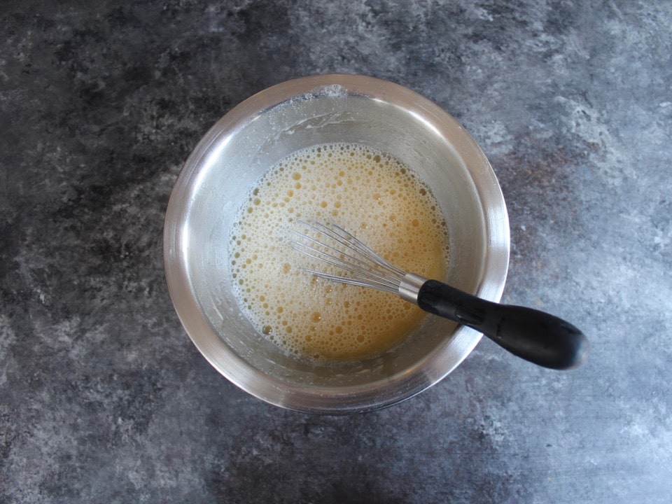 Small mixing bowl with egg, liquid and whisk in it on a concrete background.