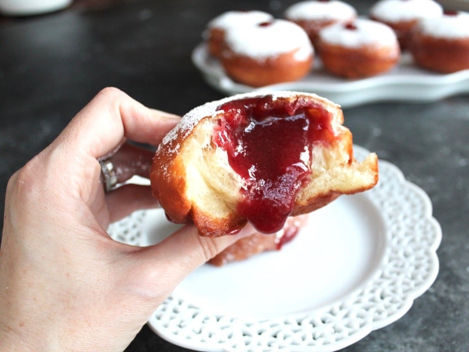 Hand holding sufganiyot that has been bitten into, revealing red jam oozing out of the center. Plate and platter of sufganiyot in background.