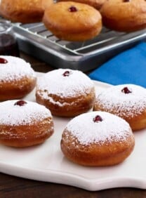 Sufganiyot on white platter dusted with sugar, more in background on cooling rack, with blue cloth towel.