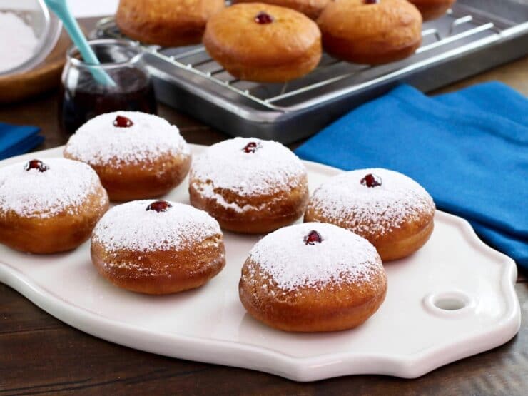 Sufganiyot on white platter dusted with sugar, more in background on cooling rack, with blue cloth towel. 