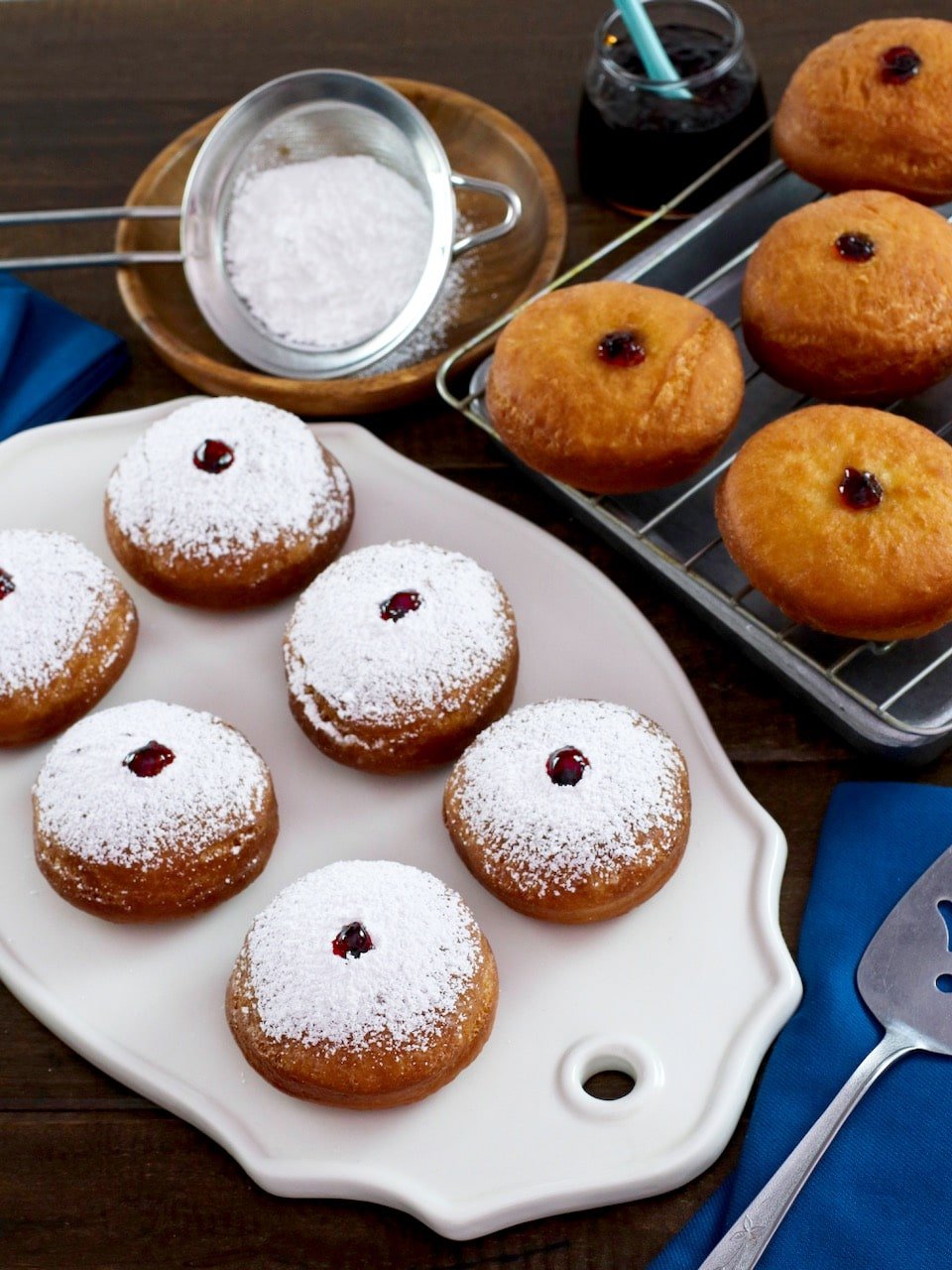 Overhead shot - sufganiyot on white platter dusted with sugar, more in background on cooling rack, with blue cloth towel. Sifter with sugar in background.