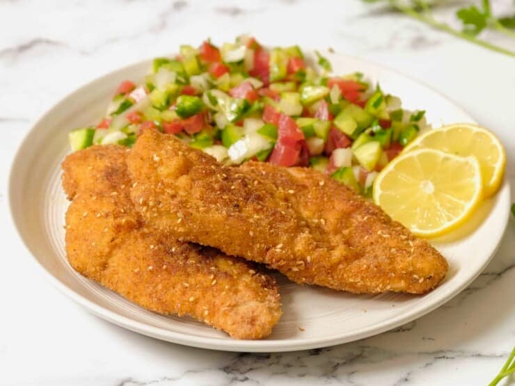 Close up - plate of chicken schnitzel, chopped salad in background with lemon, on marble counter.