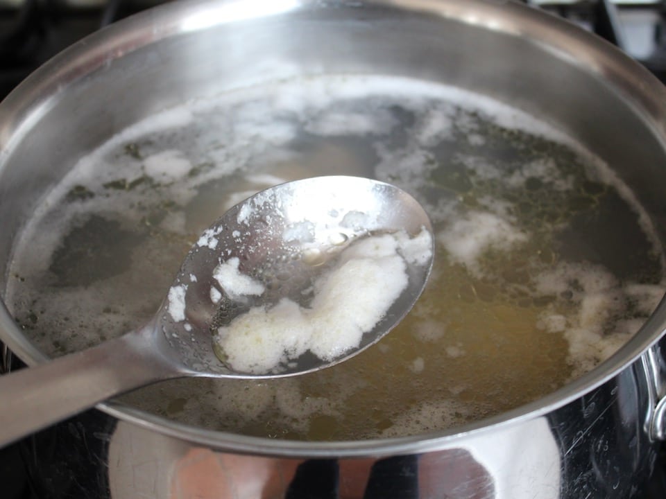 Spoon skimming foam from the surface of stock pot water.