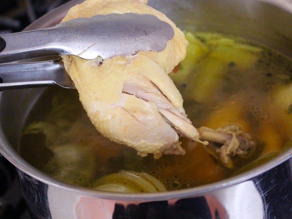 Tongs holding chicken leg up out of soup pot, tender meat falling from the bone.