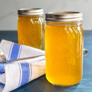 Horizontal shot of two jars of chicken stock on a blue surface with cloth napkin and soup ladling spoon.