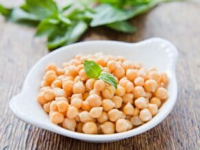White dish of cooked Mediterranean chickpeas with fresh basil garnish on a wooden table, basil in background.