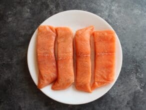 Four raw salmon fillets on a white plate seasoned with salt and pepper.