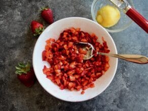 Bowl of macerating strawberries with spoon, three whole strawberries on one side, small bowl with lemon and zesting tool on the other side.