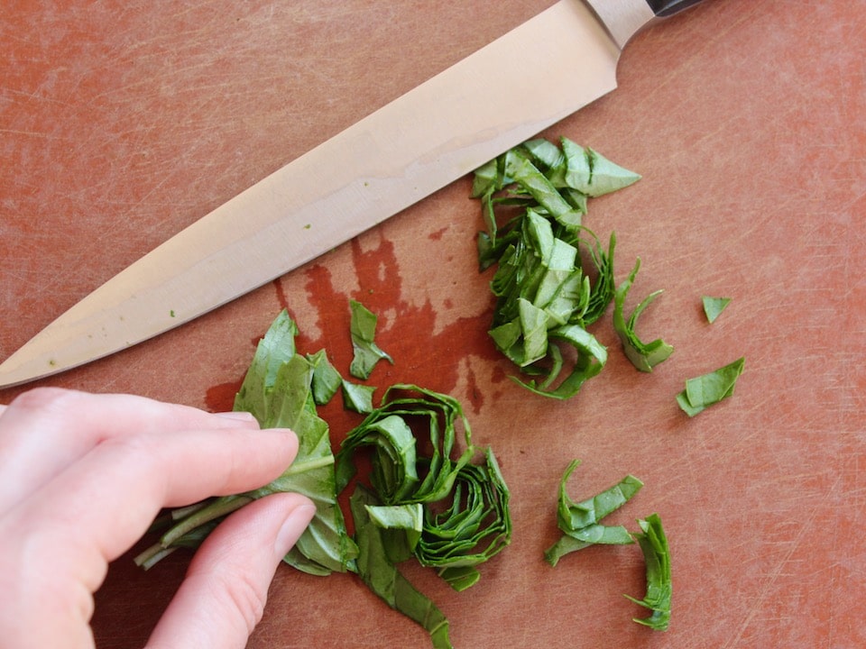 Hand holding basil leaves which have been sliced into thin chiffonade slices, knife resting on cutting board nearby.