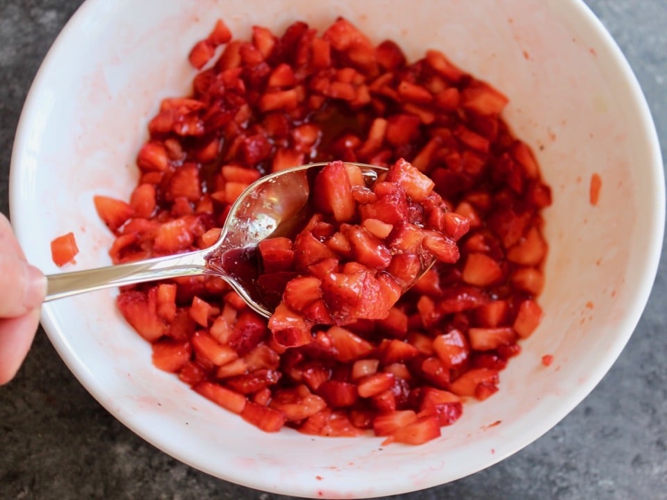 Bowl of macerating strawberries, spoon showing thickened texture in close up to camera.