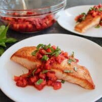 Horizontal Crop - seared salmon fillet topped with macerated strawberry balsamic sauce and fresh basil chiffonade, another plate of the same entree in background, with a glass bowl of macerating strawberries and a pile of fresh basil in the back.