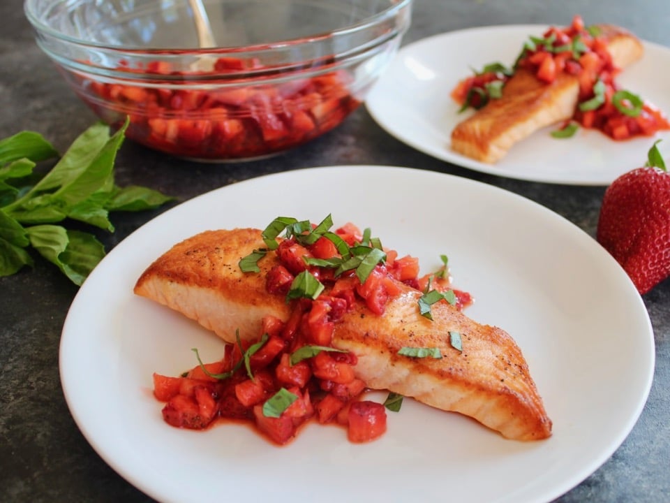 Horizontal Crop - seared salmon fillet topped with macerated strawberry balsamic sauce and fresh basil chiffonade, another plate of the same entree in background, with a glass bowl of macerating strawberries and a pile of fresh basil in the back.