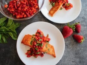 Overhead shot - plate of seared salmon topped with strawberry sauce and chopped basil, another plate nearby, bowl of macerating strawberries, fresh basil on the table with whole strawberries nearby.