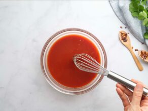 Horizontal overhead shot of a small glass dish filled with red sauce being mixed with a whisk.
