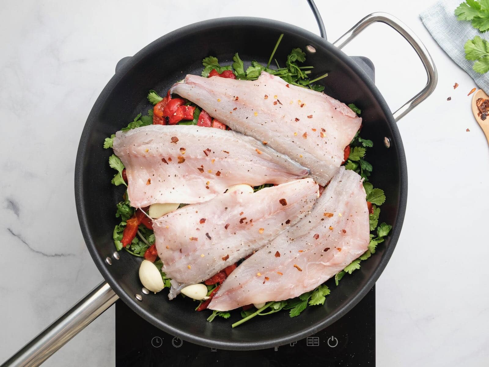 Horizontal overhead shot of a black sautee pan containing four white fish fillets laying on top of a bed of greens, red peppers, and garlic cloves.