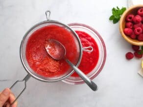 Overhead horizontal shot of a small mesh strainer containing raspberry puree. A spoon is pushing the puree through the strainer and into a small glass dish.
