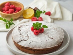 Horizontal shot of a chocolate almond flour cake topped with powdered sugar and fresh raspberries on a white plate.