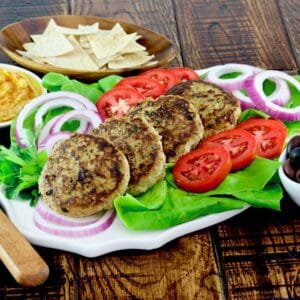 Wide horizontal shot - white plate of four turkey burgers stacked in a line garnished with lettuce, red onion and tomato slices with a dish of olives, hummus, and pita chips on the side, spatula laying nearby on wooden table.
