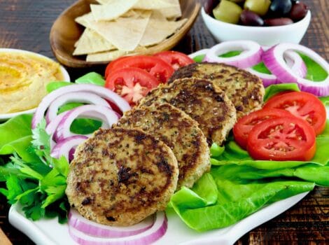 Square Crop - Plate with four grilled Mediterranean Turkey Burgers with onion slices, tomato slices and a bed of fresh green lettuce. Hummus, pita chips, and a dish of olives in the background.