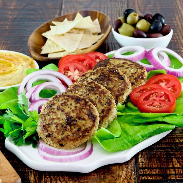 Square Crop - Plate with four grilled Mediterranean Turkey Burgers with onion slices, tomato slices and a bed of fresh green lettuce. Hummus, pita chips, and a dish of olives in the background.