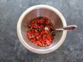 Roasted peppers and olives mixed in a steel bowl on a grey stone countertop with a spoon.