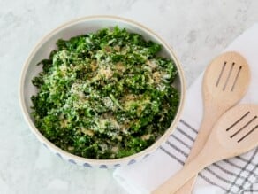 Overhead shot - large serving bowl filled with kale Caesar salad topped with creamy roasted garlic dressing, parmesan and panko breadcrumbs. Two wooden serving spoons rest on a linen napkin beside the bowl.