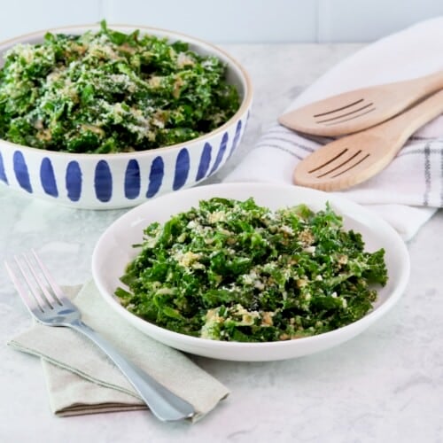 Horizontal shot - dish and serving bowls filled with kale Caesar salad on a white marble countertip. Linen napkins, metal fork, and two wooden serving spoons beside them.