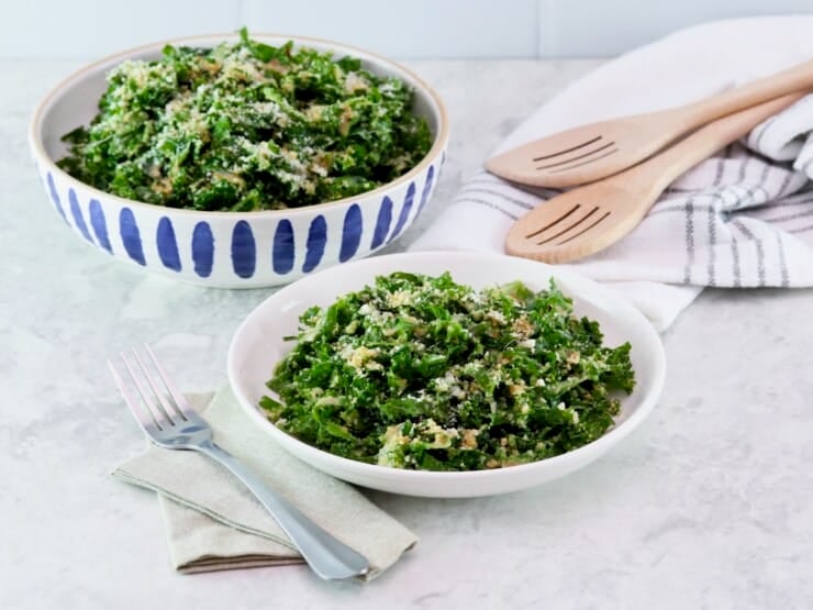 Horizontal shot - dish and serving bowls filled with kale Caesar salad on a white marble countertip. Linen napkins, metal fork, and two wooden serving spoons beside them.