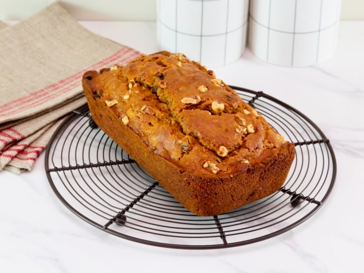 Front tilted horizontal shot - pumpkin spice cake whole loaf studded with walnuts on a circular cooling rack on a marble countertop. Linen napkin and two white ceramic jars in background.