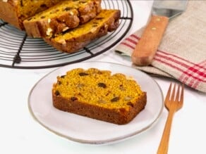 Horizontal front tilted shot - sliced pumpkin spice cake loaf on a cooling rack, with one slice on a small plate, alongside a wooden fork and linen napkin, on a white marble background.