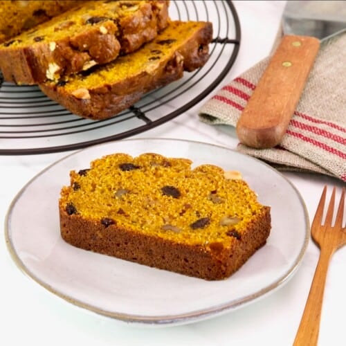 Horizontal front tilted shot - sliced pumpkin spice cake loaf on a cooling rack, with one slice on a small plate, alongside a wooden fork and linen napkin, on a white marble background.