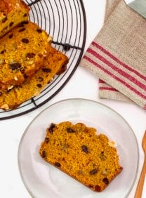 Overhead shot of sliced pumpkin spice cake loaf on a cooling rack, with one slice on a small plate, alongside a wooden fork and linen napkin, on a white marble background.
