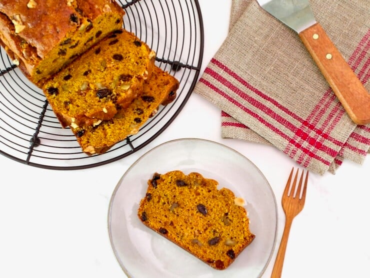 Overhead shot of sliced pumpkin spice cake loaf on a cooling rack, with one slice on a small plate, alongside a wooden fork and linen napkin, on a white marble background.