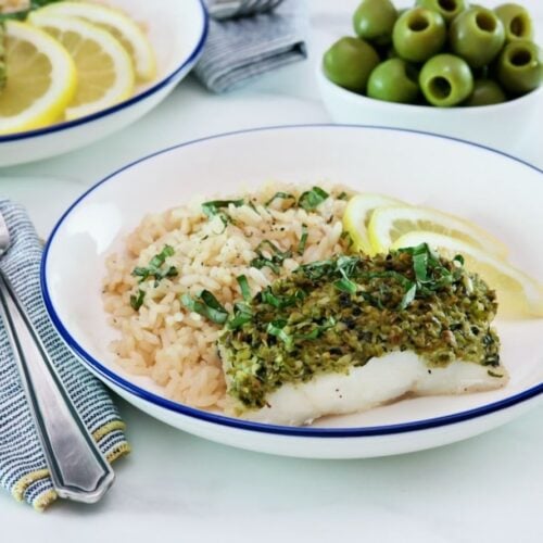 White fish fillet topped with basil tapenade in a bowl alongside couscous and lemon slices.