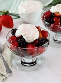 Horizontal shot of two glass dessert dishes filled with a mixture of berries and topped with whipped cream. Two whole strawberries, a dish of whipped cream, and a green tea towel are in the background.