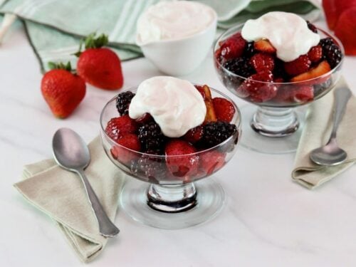 Horizontal shot of two glass dessert dishes filled with a mixture of berries and topped with whipped cream. Two whole strawberries, a dish of whipped cream, and a green tea towel are in the background.