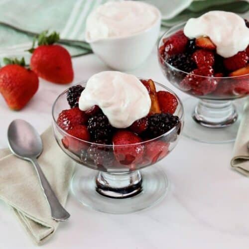 Horizontal shot of two glass dessert dishes filled with a mixture of berries and topped with whipped cream. Two whole strawberries, a dish of whipped cream, and a green tea towel are in the background.