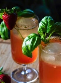Two cocktail glasses filled with a red beverage and garnished with a sprig of basil and a strawberry.
