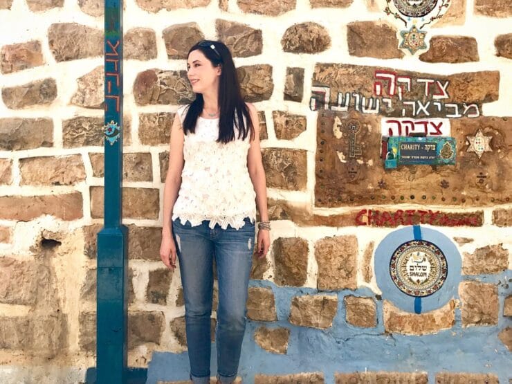 Tori Avey in Israel - Streets of Safed. Tori leans against a brick wall with Hebrew graffiti next to a light pole, wearing a white top, jeans, and sunglasses.