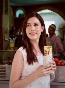 Tori Avey in front of a restaurant in Safed, Israel smiling and holding a Yemenite lachuch bread wrap. Tori wears a white top.