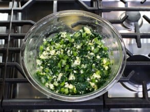 Overhead shot of a glass mixing bowl filled with spanakopita ingredients - egg, spinach, feta, onion, dill, and chives.