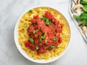 Overhead horizontal shot of a bowl egg noodles topped with chicken cacciatore in tomato sauce and garnished with parsley, on a marble countertop.