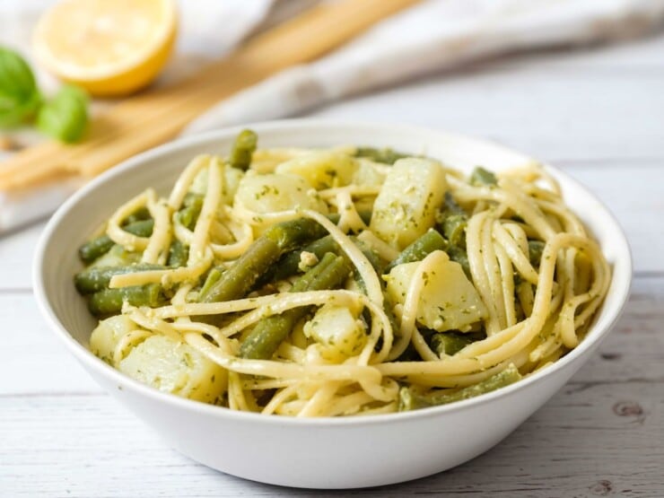 Close up of a large bowl of trenette al pesto - delicious pasta recipe with trenette or linguine pasta, potatoes, and green beans in a white bowl on a marble counter. Lemon and basil with towel in background.