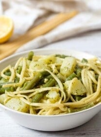 Vertical wide shot of trenette al pesto - delicious pasta recipe with trenette or linguine pasta, potatoes, and green beans in a white bowl on a marble counter. Lemon and basil with towel in background.