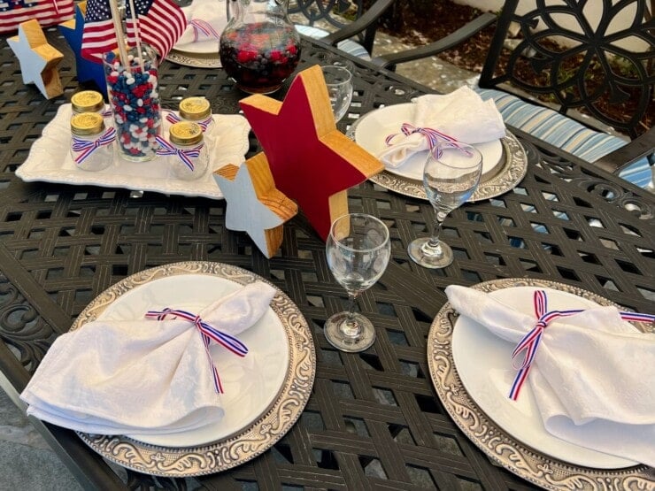 Linen napkins on white plates with red white and blue ribbon napkin rings, wine glasses, and wooden stars with a 4th of July centerpiece on a black metal outdoor table.