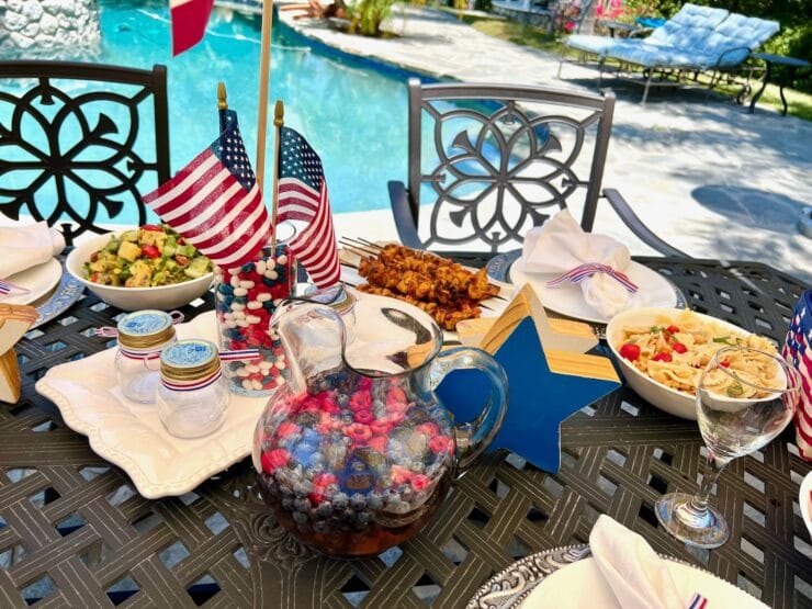 Outdoor table near a sparkling pool decorated for 4th of July. Berry sangria, pasta salad, chicken skewers, grilled salad on the table with festive 4th of July decorations and table settings. Outdoor chairs in background. 