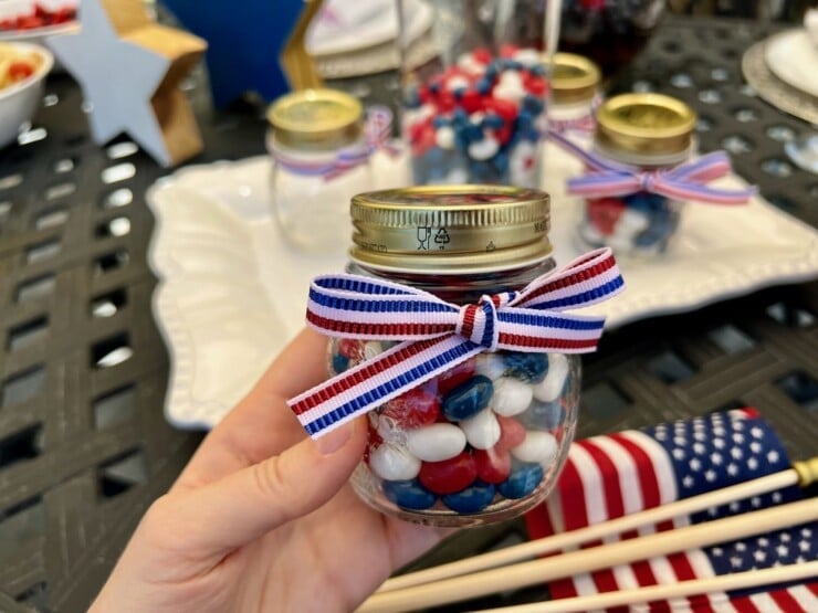 Hand holding small mason jar with red, white, and blue candies, wrapped with a small ribbon. Outdoor table in background decorated for the 4th of July with flags and other decor.