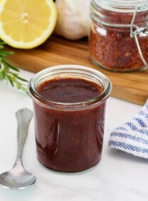 Horizontal shot of a glass jar filled with a dark red marinade, fresh herbs, lemon, and pepper flakes sit in the background.
