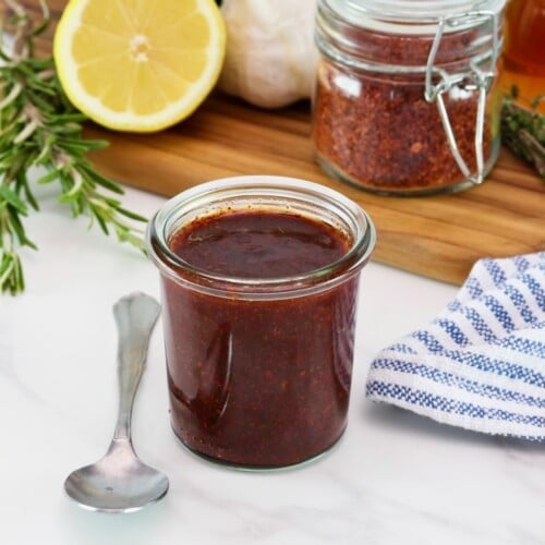 Horizontal shot of a glass jar filled with a dark red marinade, fresh herbs, lemon, and pepper flakes sit in the background.