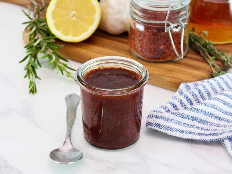 Horizontal shot of a glass jar filled with a dark red marinade, fresh herbs, lemon, and pepper flakes sit in the background.